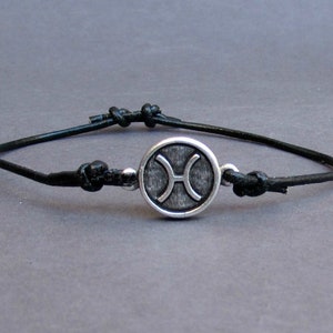 May include: A black leather bracelet with a silver charm featuring the Pisces zodiac symbol.