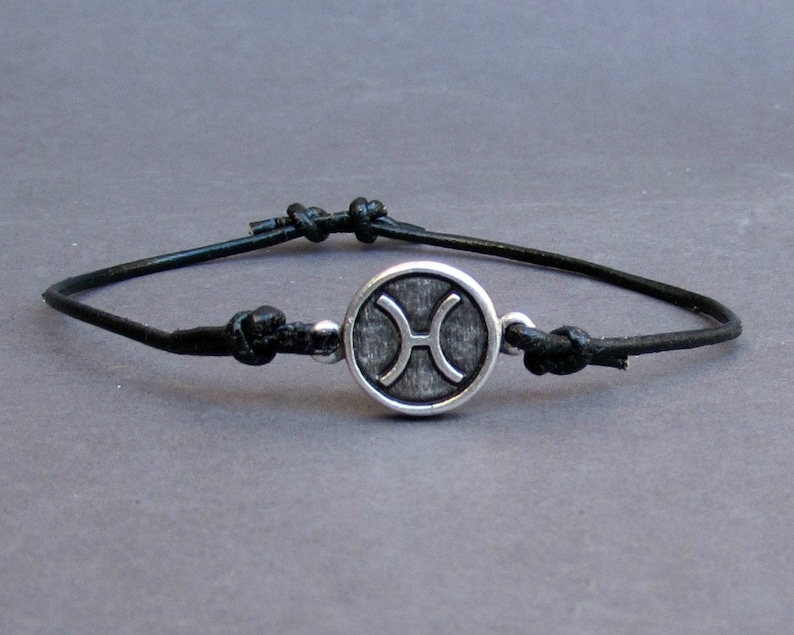 May include: A black leather bracelet with a silver charm featuring the Pisces zodiac symbol.