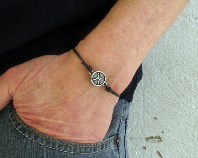 May include: A black leather bracelet with a silver charm featuring a stylized symbol. The bracelet is worn on a person's wrist.