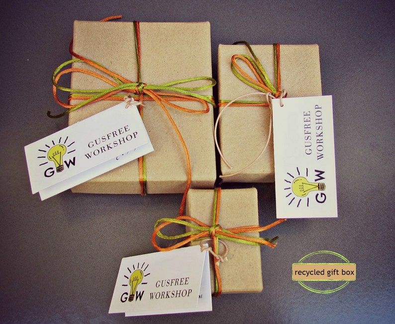 May include: Three brown cardboard gift boxes tied with orange and green ribbon. Each box has a white tag with a lightbulb graphic and the text "Gusfree Workshop" and "GW". The boxes are on a gray surface. The text "recycled gift box" is written in a gold box on the bottom right corner of the image.
