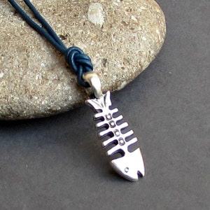 Puede incluir: Colgante de plata con forma de esqueleto de pez sobre un collar de cordón azul oscuro. El colgante tiene detalles de costillas y forma de cabeza. El cordón está anudado en la parte superior y sujeto a una pequeña cuenta plateada.