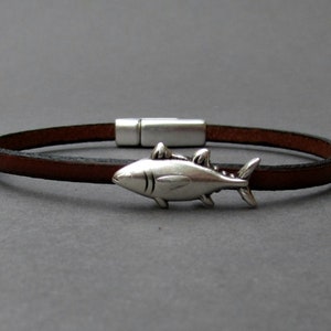 May include: A brown leather bracelet with a silver fish charm. The bracelet has a magnetic clasp.