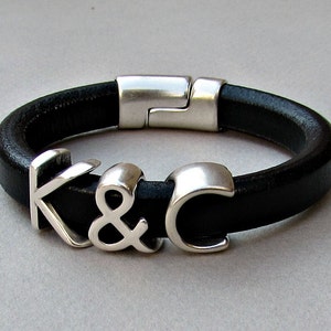 Pode incluir: Um bracelete de couro preto com fecho prateado e pingentes prateados que soletram "K & C".