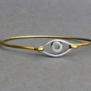 Può includere: Un braccialetto dorato con un ciondolo a forma di occhio d'argento. Il ciondolo a forma di occhio ha un centro rotondo ed è circondato da una cornice d'argento.