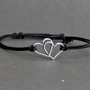 Könnte beinhalten: Ein schwarzes Lederarmband mit einem silbernen Doppelherzen-Anhänger.