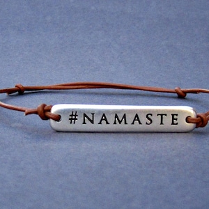 Puede incluir: Un brazalete de cuero marr&oacute;n con un dije de plata que dice "#NAMASTE".