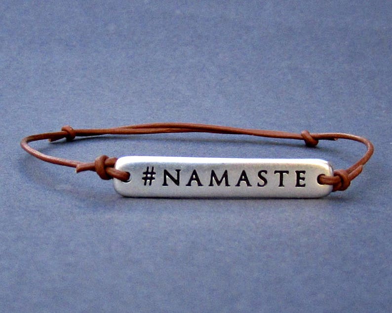 Puede incluir: Un brazalete de cuero marr&oacute;n con un dije de plata que dice "#NAMASTE".