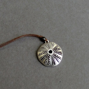 Puede incluir: Un collar de plata con un colgante redondo con forma de erizo de mar. El colgante tiene una superficie texturizada con pequeños bultos y un centro negro.