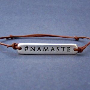 Puede incluir: Un brazalete de cuero marr&oacute;n con un dije de plata que dice "#NAMASTE".