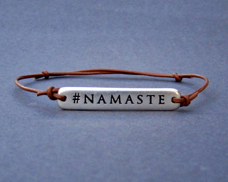 Puede incluir: Un brazalete de cuero marr&oacute;n con un dije de plata que dice "#NAMASTE".