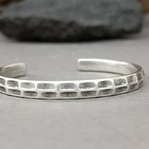 May include: A silver cuff bracelet with a textured, brick-like pattern. The open design allows for easy wear. The bracelet is displayed on a grey surface, showcasing its metallic sheen. A simple, yet stylish piece of jewellery.
