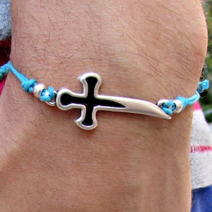 May include: A turquoise cord bracelet with a silver sword-shaped cross charm. The cross is black in the center.