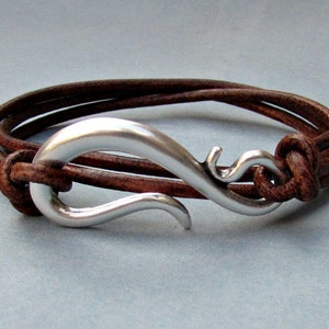 May include: A brown leather bracelet with a silver hook clasp. The bracelet is made of two strands of leather and the hook is a simple, elegant design.
