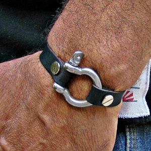 Può includere: Un braccialetto in pelle nera con una chiusura in metallo argentato. La chiusura è un anello rotondo con una piccola vite sul lato.