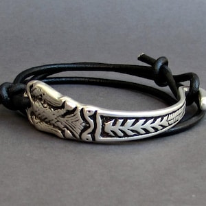 May include: A silver bracelet with a detailed design of a bird and leaves. The bracelet is attached to a black leather strap with a knot.