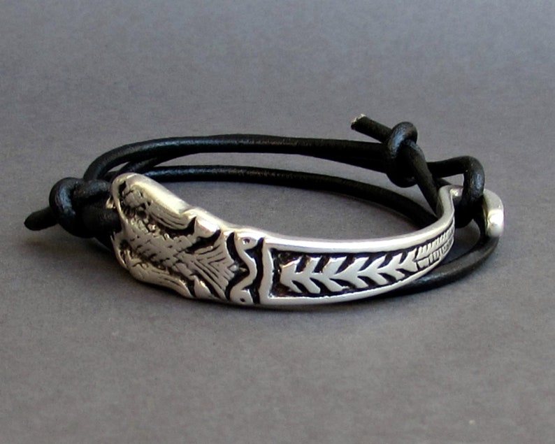 May include: A silver bracelet with a detailed design of a bird and leaves. The bracelet is attached to a black leather strap with a knot.