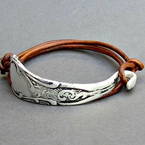 May include: A brown leather bracelet with a silver spoon charm. The spoon has an ornate design and is attached to the leather strap with a silver clasp.