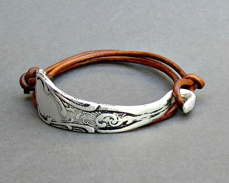 May include: A brown leather bracelet with a silver spoon charm. The spoon has an ornate design and is attached to the leather strap with a silver clasp.