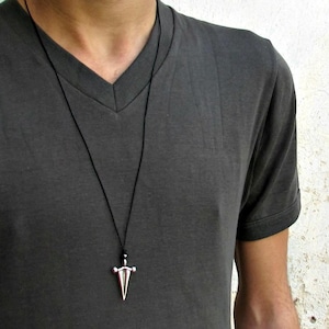 May include: A silver pendant in the shape of a sword hangs from a black cord necklace. The pendant is worn over a dark gray V-neck t-shirt. The background is a white wall.