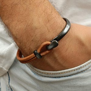 Può includere: Un bracciale a polsino in metallo scuro con un cinturino in pelle marrone. Il bracciale è indossato su un polso, mostrando il contrasto tra il metallo e la pelle. Il design è semplice e moderno, adatto all'uso quotidiano.