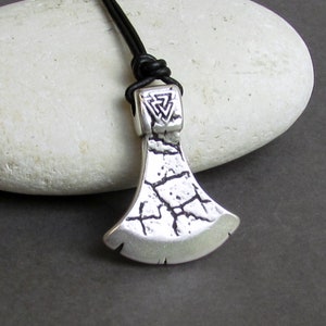 May include: Silver axe pendant with a textured surface and a triangular knot design. The pendant is on a black leather cord.