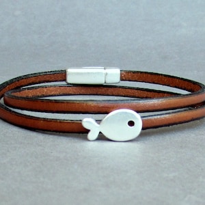Könnte beinhalten: Ein braunes Lederarmband mit einem silbernen Fisch-Anhänger. Das Armband ist doppelt gewickelt und hat einen Magnetverschluss.