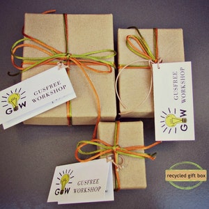 Puede incluir: Tres cajas de regalo de cart&oacute;n marr&oacute;n atadas con cinta naranja y verde. Cada caja tiene una etiqueta blanca con un gr&aacute;fico de bombilla y el texto "Gusfree Workshop" y "GW". Las cajas est&aacute;n sobre una superficie gris. El texto "caja de regalo reciclada" est&aacute; escrito en una caja dorada en la esquina inferior derecha de la imagen.