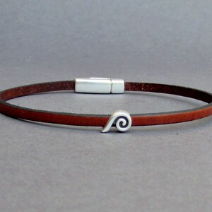Puede incluir: Un brazalete de cuero marrón con un cierre magnético plateado y un dije de espiral plateado.