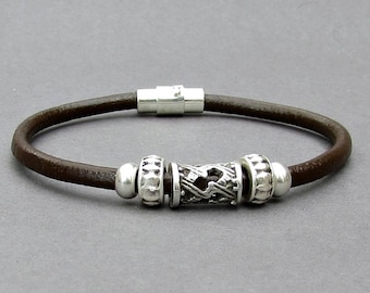 May include: A brown leather bracelet with a silver magnetic clasp and silver bead accents. The center bead features a geometric design.