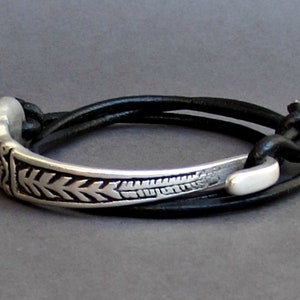 May include: A silver bracelet with a decorative design and a black leather strap. The strap is adjustable and can be worn on the wrist or ankle.
