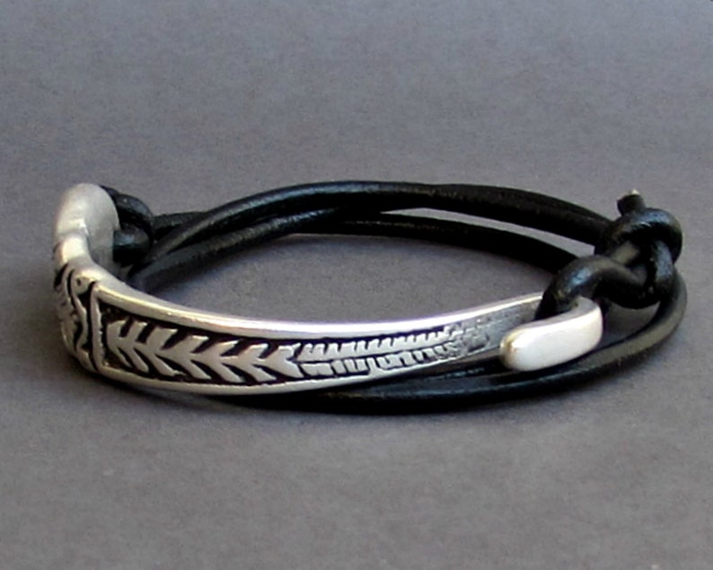 May include: A silver bracelet with a decorative design and a black leather strap. The strap is adjustable and can be worn on the wrist or ankle.