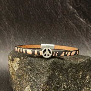 Puede incluir: Un brazalete de cuero negro y marrón con un dije de plata en forma de signo de paz.