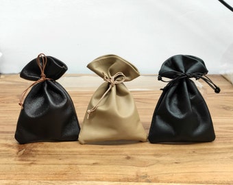 Pode incluir: Três bolsas de couro com cordão em preto, bege e preto. As bolsas são fechadas com um cordão de couro. As bolsas estão sobre uma superfície de madeira.