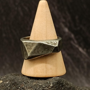 Può includere: Un anello d'argento con un design geometrico sfaccettato. L'anello è esposto su un porta anelli in legno.