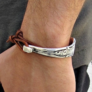 May include: A brown leather bracelet with a silver metal cuff. The cuff has a decorative design.