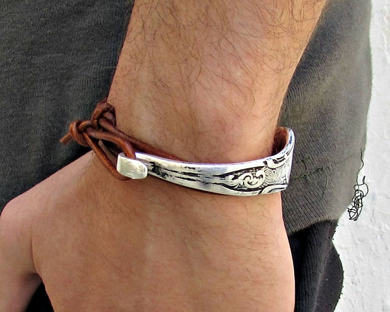 May include: A brown leather bracelet with a silver metal cuff. The cuff has a decorative design.