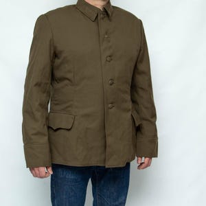 Könnte beinhalten: Olivgrüne Jacke mit Knopfleiste, Kragen und zwei Vordertaschen. Die Jacke hat lange Ärmel und wird mit einer blauen Jeans getragen. Die Jacke ist einfarbig und scheint aus einem strapazierfähigen Stoff zu sein.