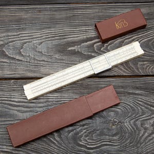 May include: A beige slide rule with a clear plastic cursor featuring black numerical markings and scales. The instrument is accompanied by a brown rectangular storage case and a separate brown lid with the gold-coloured logo text K&E.