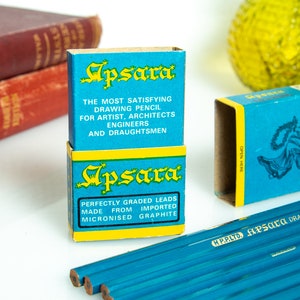 May include: Two blue and yellow Apsara drawing pencil boxes are stacked, with text that reads "THE MOST SATISFYING DRAWING PENCIL" and "PERFECTLY GRADED LEADS." Several blue pencils are visible. The pencils are for artists, architects, engineers, and draughtsmen.