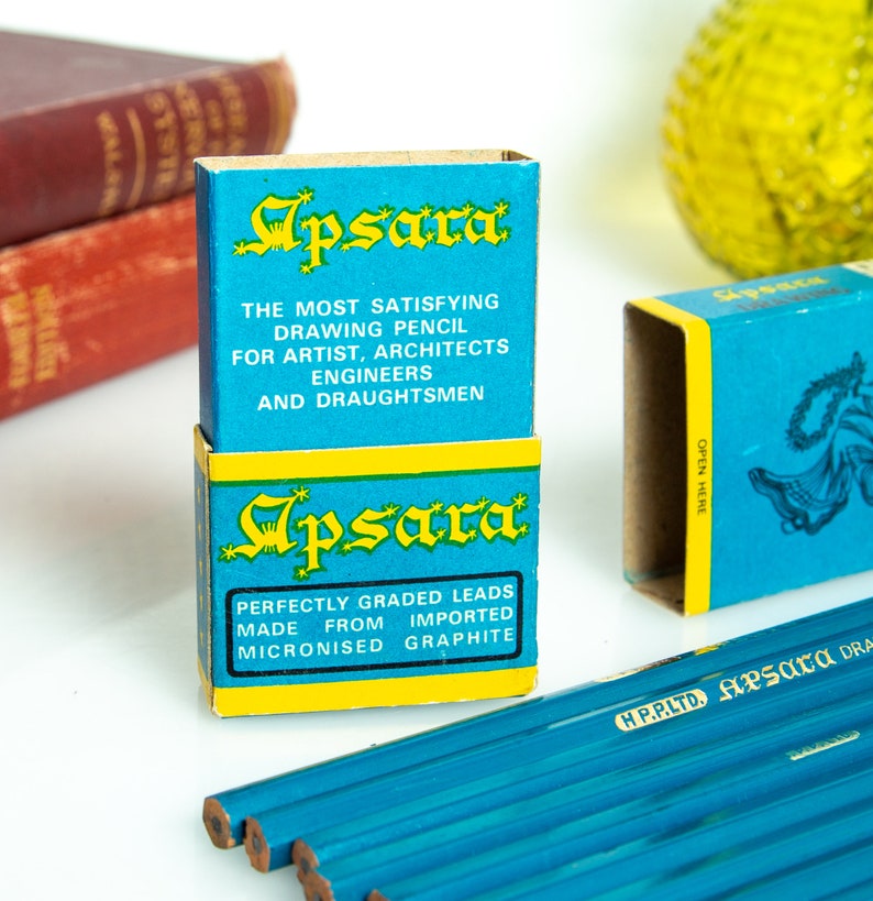 May include: Two blue and yellow Apsara drawing pencil boxes are stacked, with text that reads "THE MOST SATISFYING DRAWING PENCIL" and "PERFECTLY GRADED LEADS." Several blue pencils are visible. The pencils are for artists, architects, engineers, and draughtsmen.