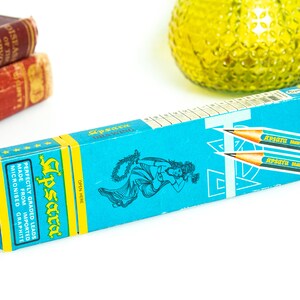 May include: A turquoise pencil box with yellow accents and the brand name "Apsara" in a stylized font. The box features illustrations of a woman and pencils, with text indicating the pencils are made from imported micronized graphite.