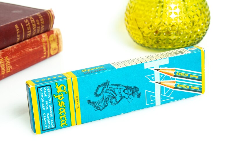 May include: A turquoise pencil box with yellow accents and the brand name "Apsara" in a stylized font. The box features illustrations of a woman and pencils, with text indicating the pencils are made from imported micronized graphite.
