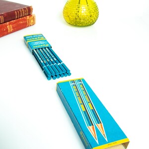 May include: A box of blue pencils with a yellow and blue design, and the word "Gypser" printed on the box. The pencils are arranged on a white surface, with a yellow glass vase and two red books in the background.
