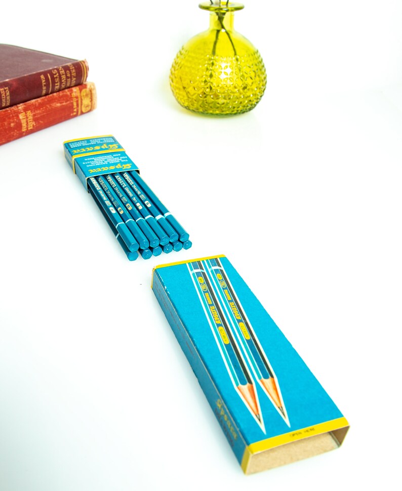 May include: A box of blue pencils with a yellow and blue design, and the word "Gypser" printed on the box. The pencils are arranged on a white surface, with a yellow glass vase and two red books in the background.