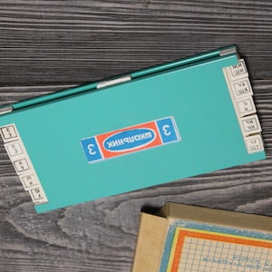 Puede incluir: Un organizador de útiles escolares vintage de color turquesa con pestañas etiquetadas. El organizador tiene forma rectangular con tapa abatible y una etiqueta que dice "ШКОЛЬНИК" en ruso. Un cuaderno parcialmente visible está en primer plano.