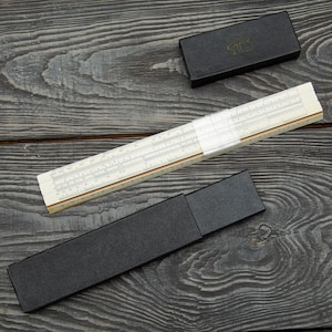 May include: A white plastic slide rule with a clear sliding cursor and multiple printed numerical scales. The instrument is accompanied by a long black rectangular storage case and a smaller black rectangular box with a gold logo on the lid.