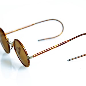 May include: A pair of round, brown tortoise shell sunglasses with brown lenses. The sunglasses have thin metal arms and are folded in half.