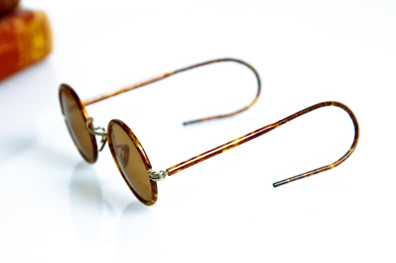 May include: A pair of round, brown tortoise shell sunglasses with brown lenses. The sunglasses have thin metal arms and are folded in half.