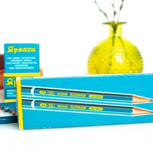 May include: A box of Apsara drawing pencils in a teal and yellow box. The pencils are blue with a silver band and a pink eraser. The box has the text "Apsara" in teal and white. A yellow vase and red books are in the background.