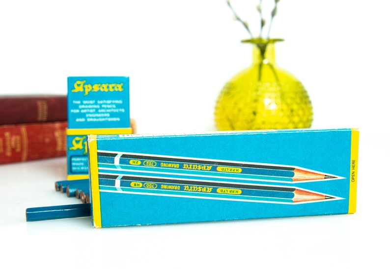 May include: A box of Apsara drawing pencils in a teal and yellow box. The pencils are blue with a silver band and a pink eraser. The box has the text "Apsara" in teal and white. A yellow vase and red books are in the background.